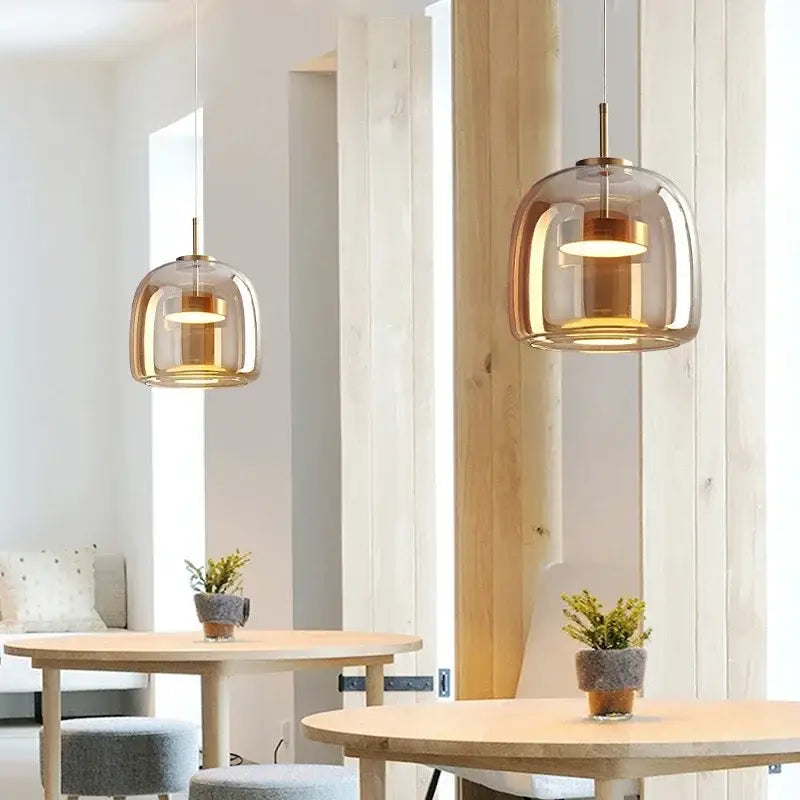 Two modern amber glass led pendant chandeliers with brass accents hang above wooden dining tables