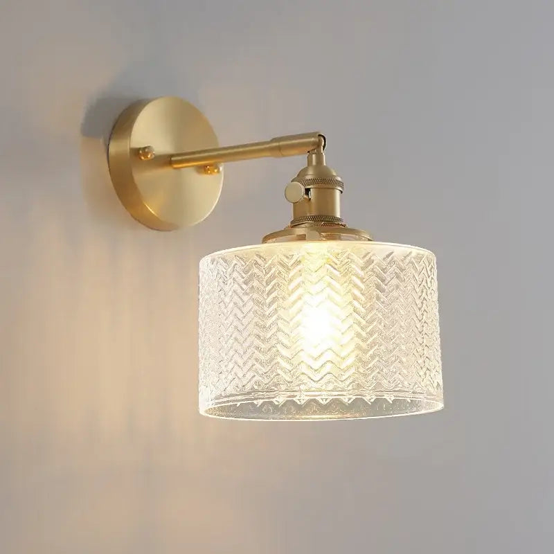 A warm-toned brass wall lamp with a cylindrical green glass led shade featuring an embossed chevron pattern