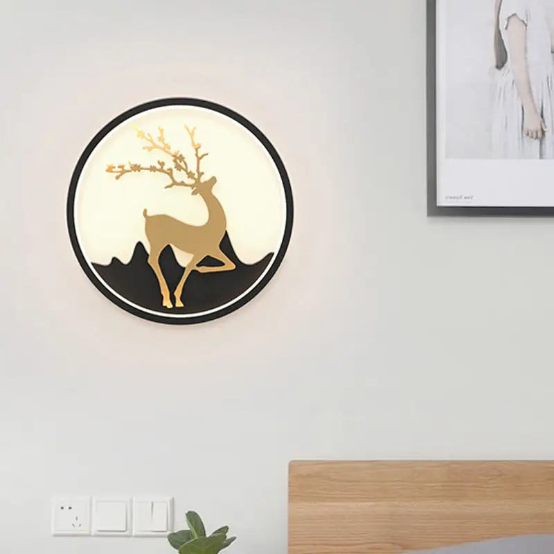 Circular wall-mounted light fixture featuring a gold deer silhouette with antlers against a black mountain backdrop, encased in a sleek black frame.
