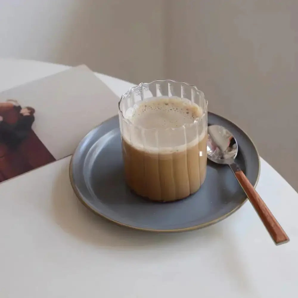 A clear fluted glass ripple cup with nordic design filled with creamy coffee on a matte gray plate with gold leaf spoon