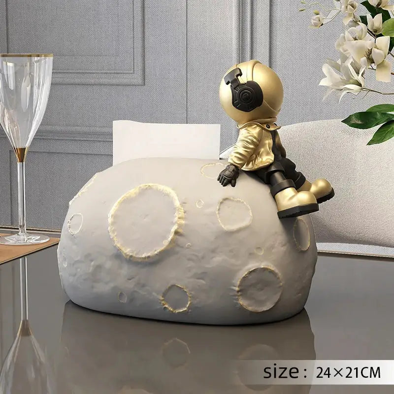 Gold astronaut figurine sitting atop a textured gray moon tissue box holder with nordic white ceramic bear statues decor