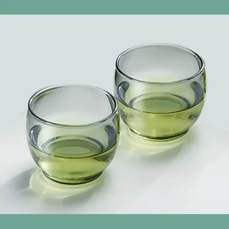 Two clear olive green glass tea cups filled with light green liquid in a decorbites glass tea cup set for kung fu tea