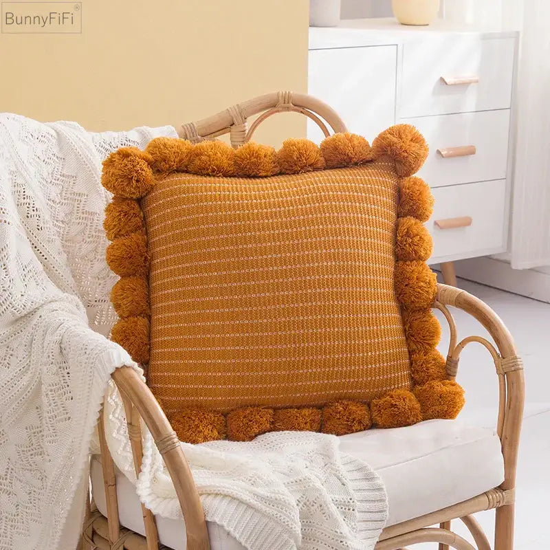Warm amber-colored woven ball cushion cover with textured knit pattern and fluffy pom-pom trim for living room decor