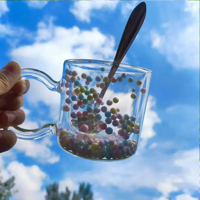 A double wall glass cup with pastel heart bubble quicksand spheres and a spoon against a bright blue sky