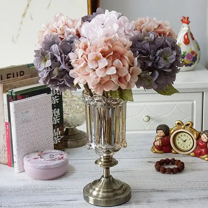 A tall clear glass vase with antique brass base holding a pastel decorbites hydrangea branch silk flower bouquet