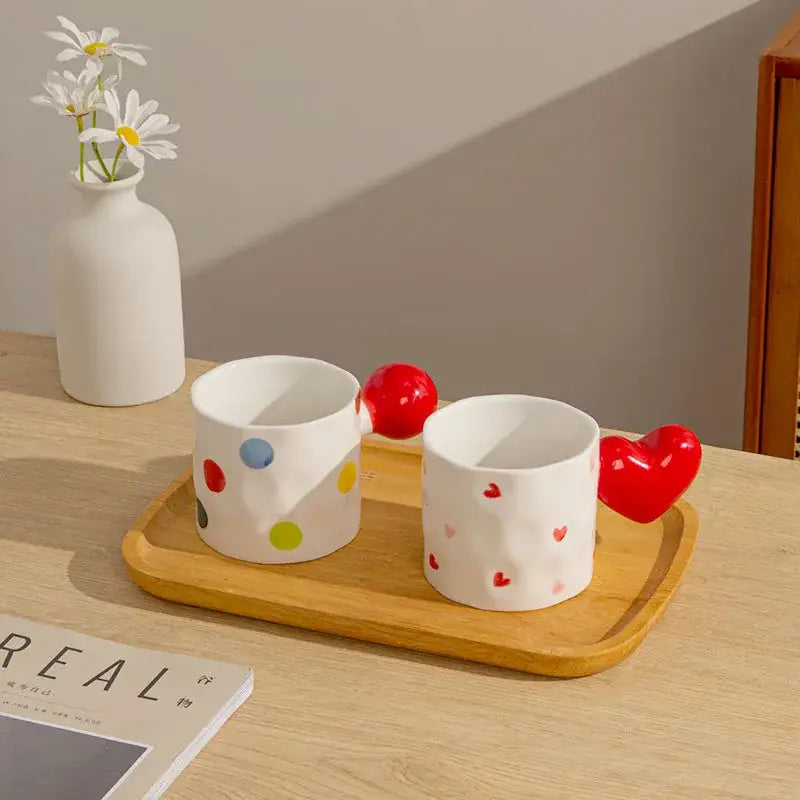 Two white ceramic mugs with colorful polka dots and red handles feature decorbites heart design on a wooden tray