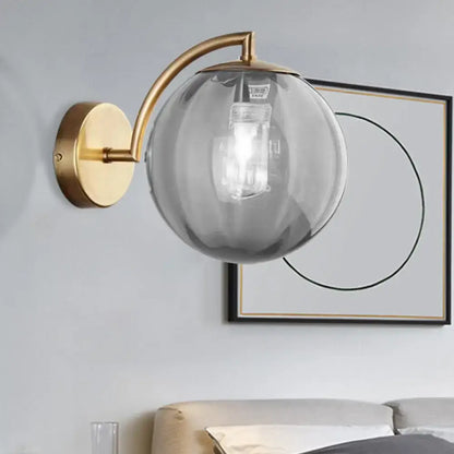 A modern brass armed ball sconce with a brushed finish and amber smoked glass globe for bedroom wall lighting