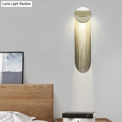 Postmodern gold-toned wall light fixture with elongated oval design and cascading vertical metal strands for bedroom use