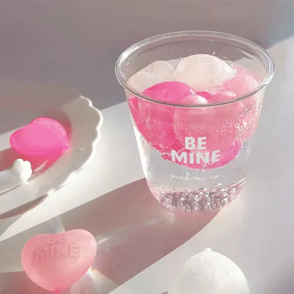A clear plastic cup with pink and white ice cubes made from purple heart ice cube mold with be mine text