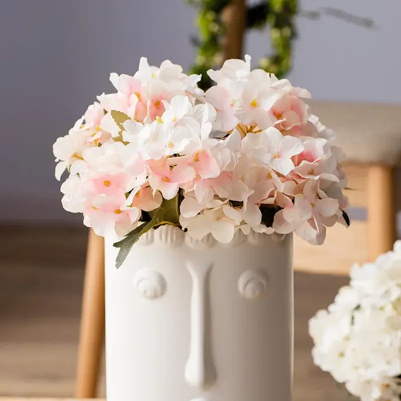 A white ceramic vase with face design holds a realistic pink and white decorbites hydrangea branch decor bouquet