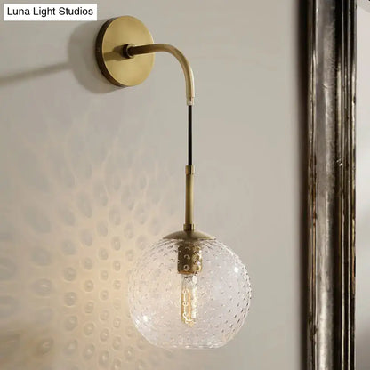 A brass wall sconce with curved arm and clear textured glass globe for pink yellow blue retro bedroom lighting