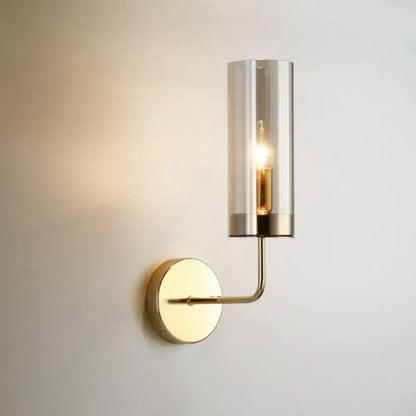 A sleek brass wall mount lamp with a cylindrical smoked glass shade and exposed filament bulb for living room use