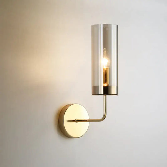 A sleek brass wall mount lamp with a cylindrical smoked glass shade and exposed filament bulb for living room use