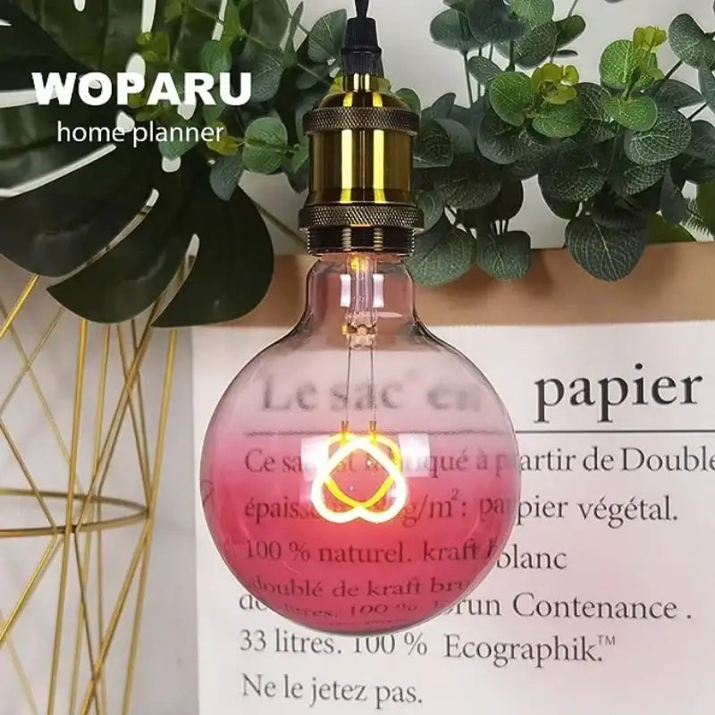 A glowing pink led filament heart light bulb with brass edison base hangs above a paper bag label decorbites gradient pink