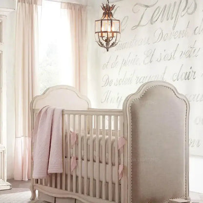 A soft beige upholstered crib with curved headboard and pink accents paired with decorbites grey gold metal cage chandelier