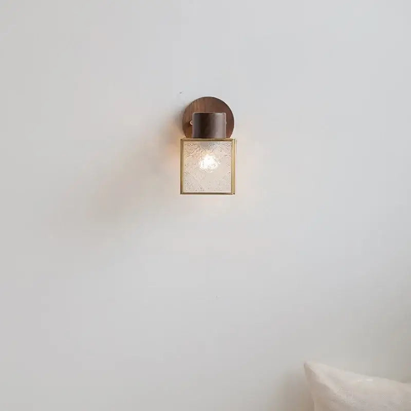 A rustic walnut wood and brass wall sconce with clear glass square shade and circular backplate for bedroom use