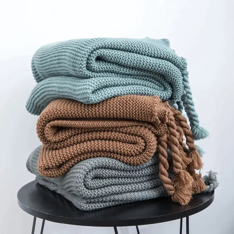 Stack of decorbites knit throw blankets in muted teal brown and gray with braided tassels for cozy decor