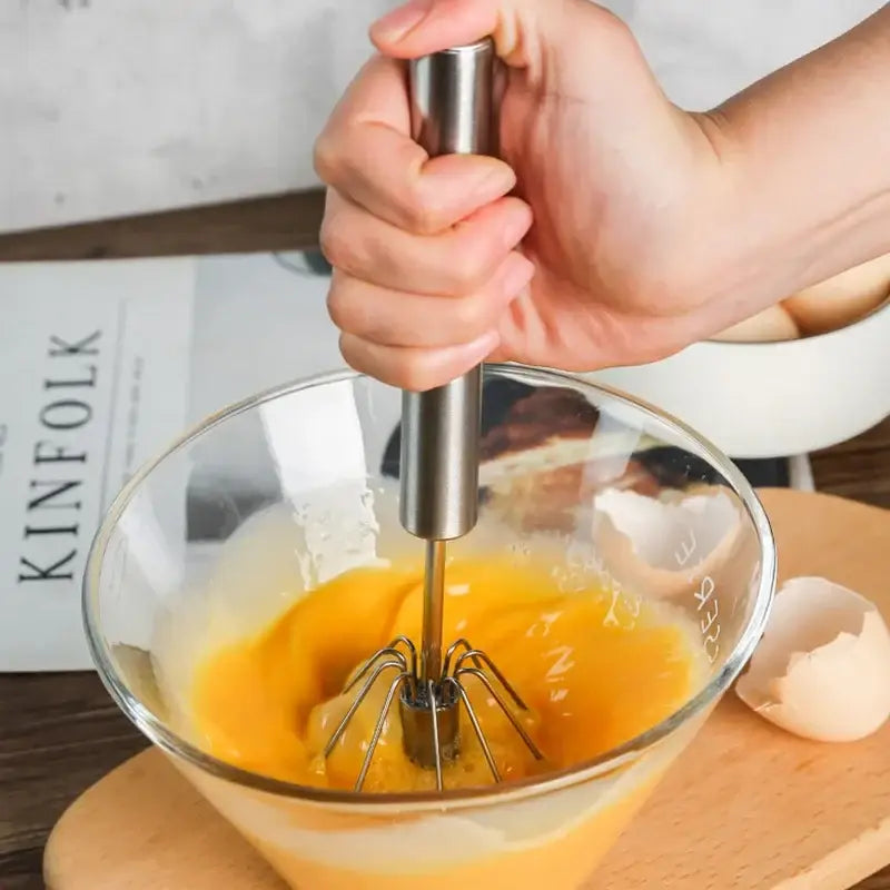 A sleek stainless steel hand pressure egg beater with polished handle whipping eggs in a clear glass bowl