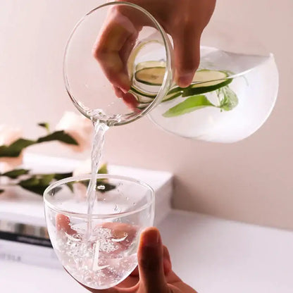 Clear teal hourglass glass tea infuser water bottle pouring cucumber mint water into matching glass carafe