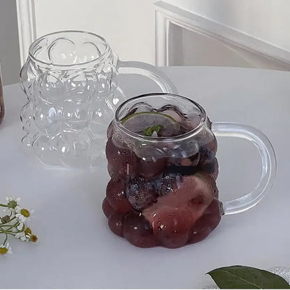 Clear glass drinking set featuring a textured grape cluster mug with transparent handle and gold spoon for dining room use