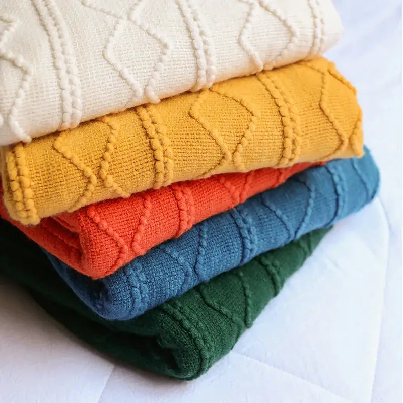 Stack of five colorful textured diamond knit throw blankets in cream, mustard, coral, teal, and forest green by decorbites cozy knit