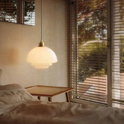 A tiered cream glass pendant light with layered opal shade and brass hardware hangs above a bedside table