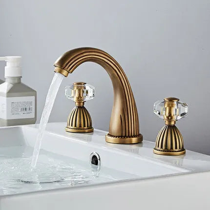 Antique brass Tuqiu gold basin faucet with crystal knob handles and curved spout flowing cold water into white sink basin