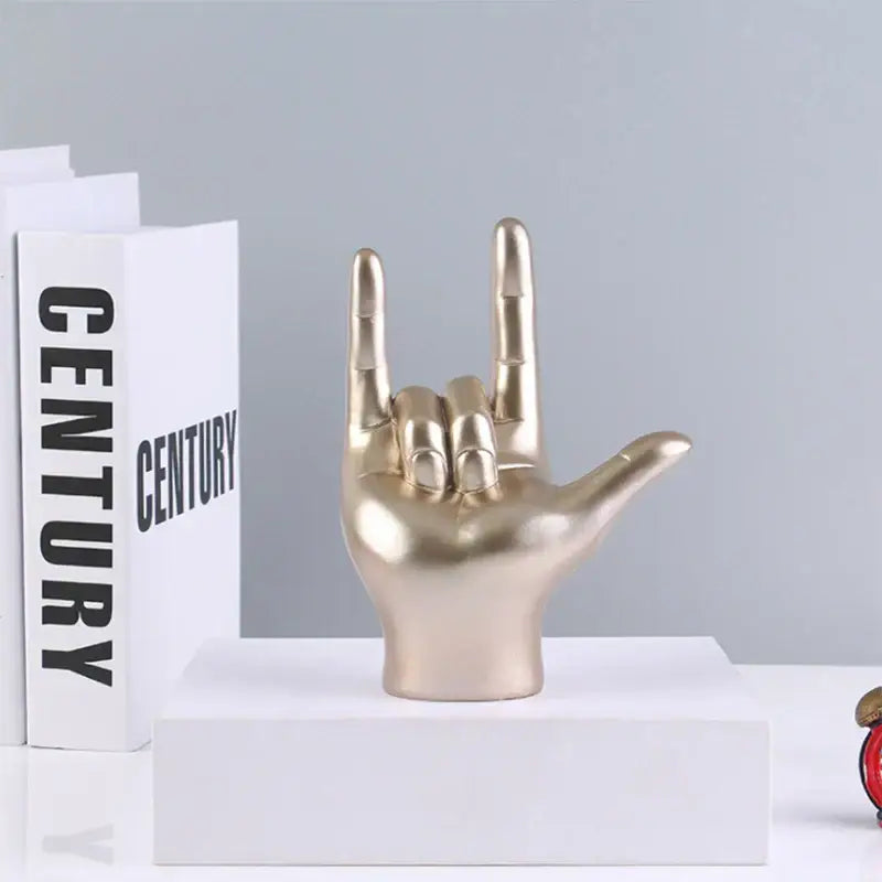Gold-colored resin decorbites abstract hand love sculpture in a rock gesture displayed on a white pedestal for living room decor