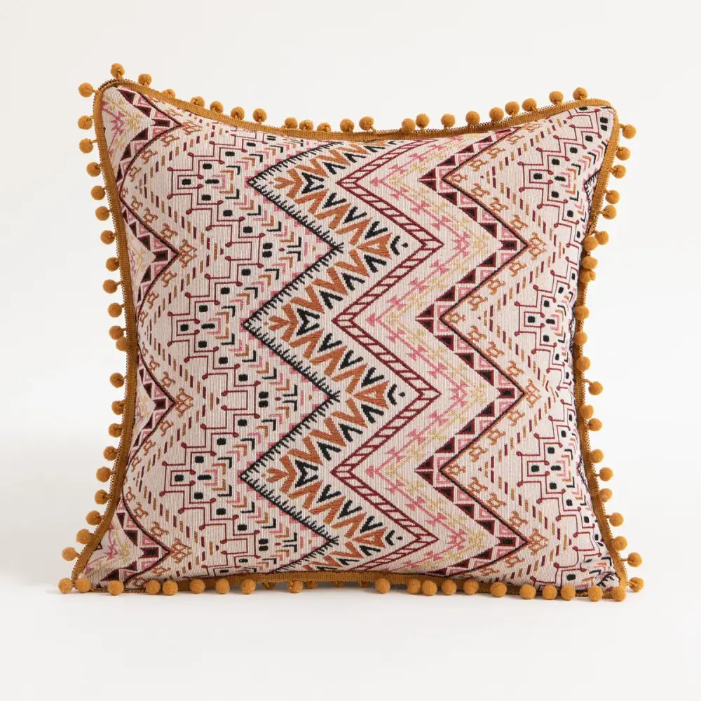 A square pillow cover with beige base and geometric rust black patterns accented by mustard pom poms for ethnic boho decorbites throw decor