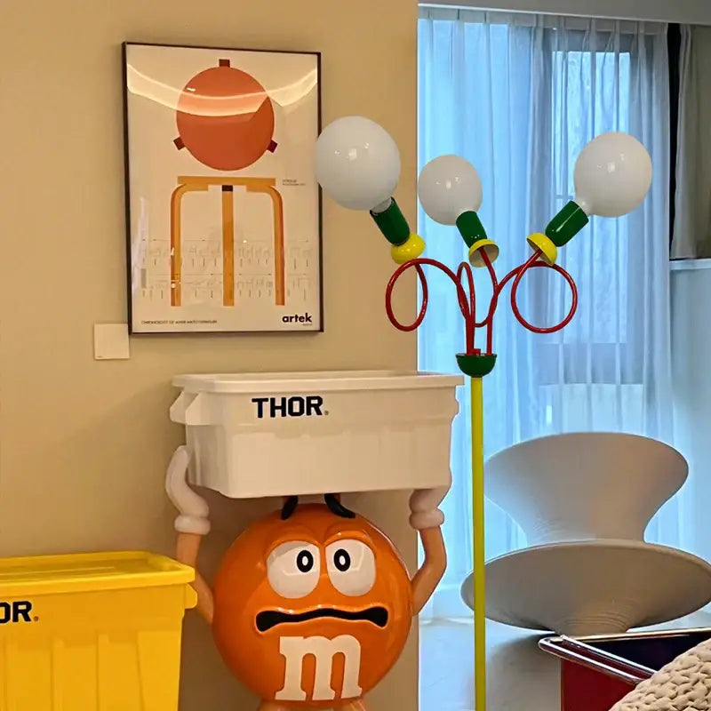 Orange m&m’s character figurine with white gloves alongside vibrant red and yellow decorbites art led floor lamp for living room decor