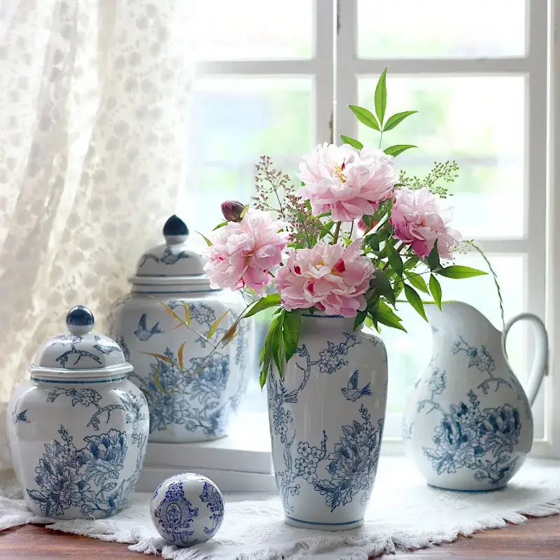 A tall white porcelain vase with intricate blue floral and bird patterns holds pink peonies for living room decor