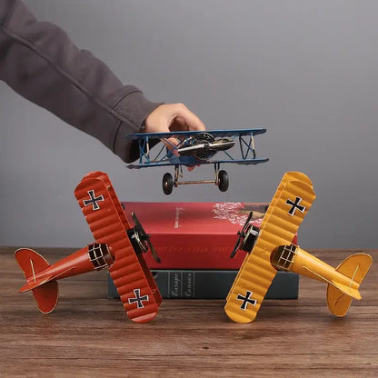 A hand holds a blue metal airplane figurine with black wheels above vintage red and yellow decorbites iron airplane models on books
