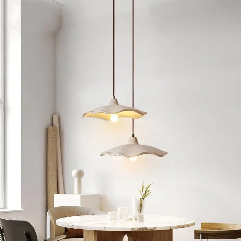 Two elegant off-white ceramic pendant lights with scalloped edges hang above a marble table with natural wood cords
