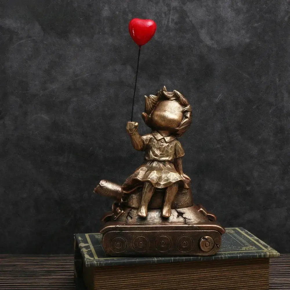 Bronze-colored resin statue of a balloon girl with curly hair sitting on a tank holding a red heart balloon for decor