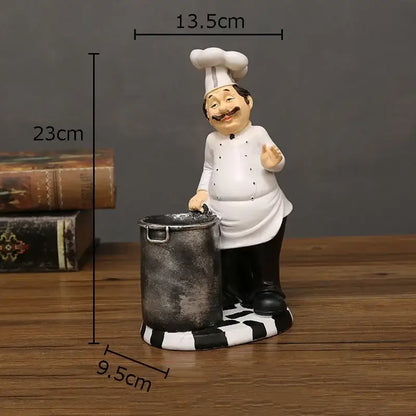 A whimsical white bearded chef resin figure with black mustache and tall hat beside a rustic metallic pot