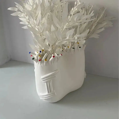 White ceramic flower pot vase shaped like coral shapes with stylized head holding paper leaves and colorful pencil tips