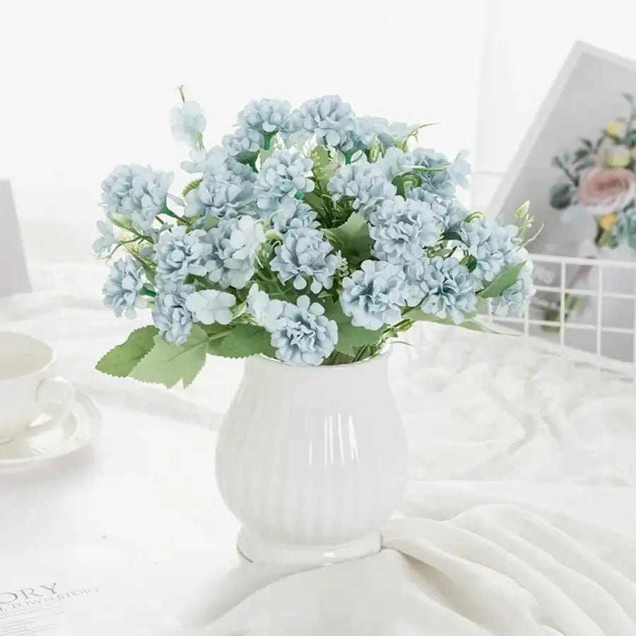 A delicate light blue artificial hydrangea bouquet with green leaves in a white ribbed ceramic vase for decorbites decor