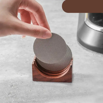 A hand holds circular metallic filter screens on a dark brown wooden espresso filter screen holder with gray coasters
